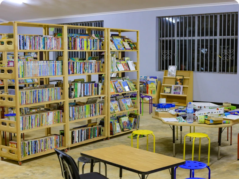 Room filled with lots of books on the shelves, tables and chairs, stools, a desk, a chair, a lamp